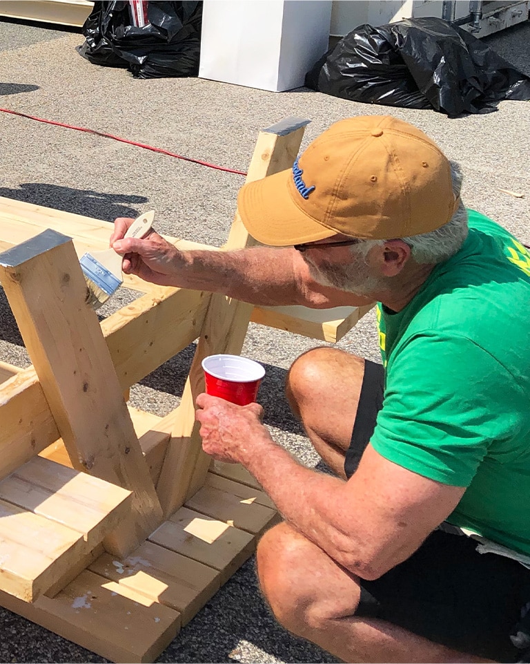 image of a man wearing a green shirt and a yellow hat crouched down staining a picnic bench